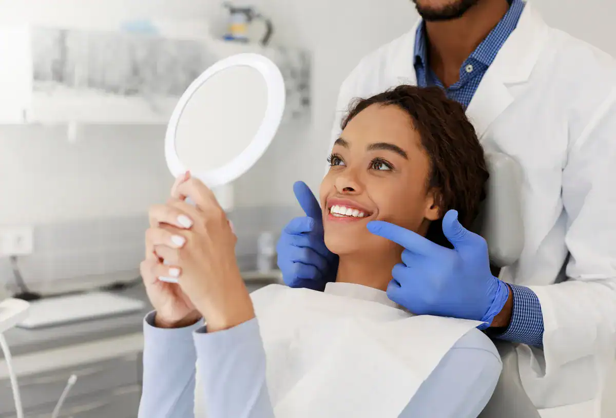 A smiling woman at the dentist's office holding a mirror for dental checkup.
